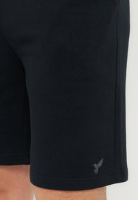 Μαύρα σορτς ακριβώς πάνω από το γόνατο με ένα μικρό λογότυπο κολιμπρί κοντά στο τελείωμα στο δεξί πόδι, φορεμένα από ένα άτομο με ορατά τα κάτω πόδια.