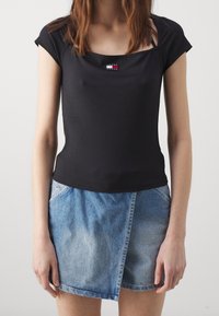 Svart kortärmad topp med fyrkantig halsringning och en liten märkespatch. Parat med en denimwrapkjol i ljusblått, som har en ficka.