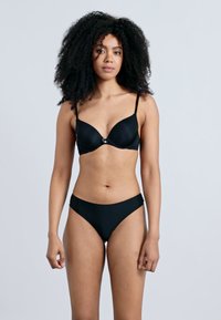 Melns krūšturis ar stīpu, gluda auduma, plānām regulējamām lencītēm un nelielu dekoratīvu priekšpuses aizdari, kopā ar melnām bikini biksītēm.