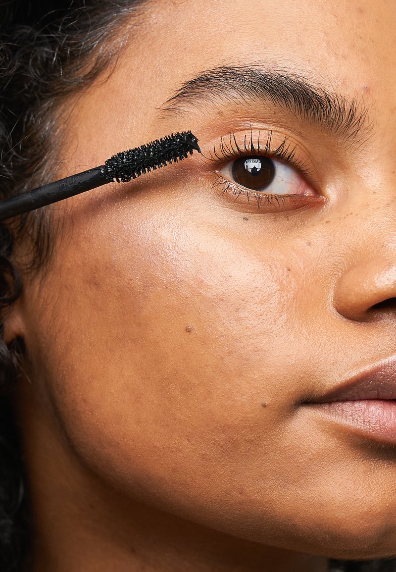 Zwarte mascara wordt met een applicator op donkere wimpers aangebracht, waardoor een close-up van een bruine oog, een egale huid en een natuurlijke wenkbrauw wordt benadrukt.