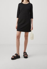 Femme portant une robe noire à manches trois-quarts, des sandales noires épaisses, tenant un petit sac à main blanc perlé et portant un collier floral.