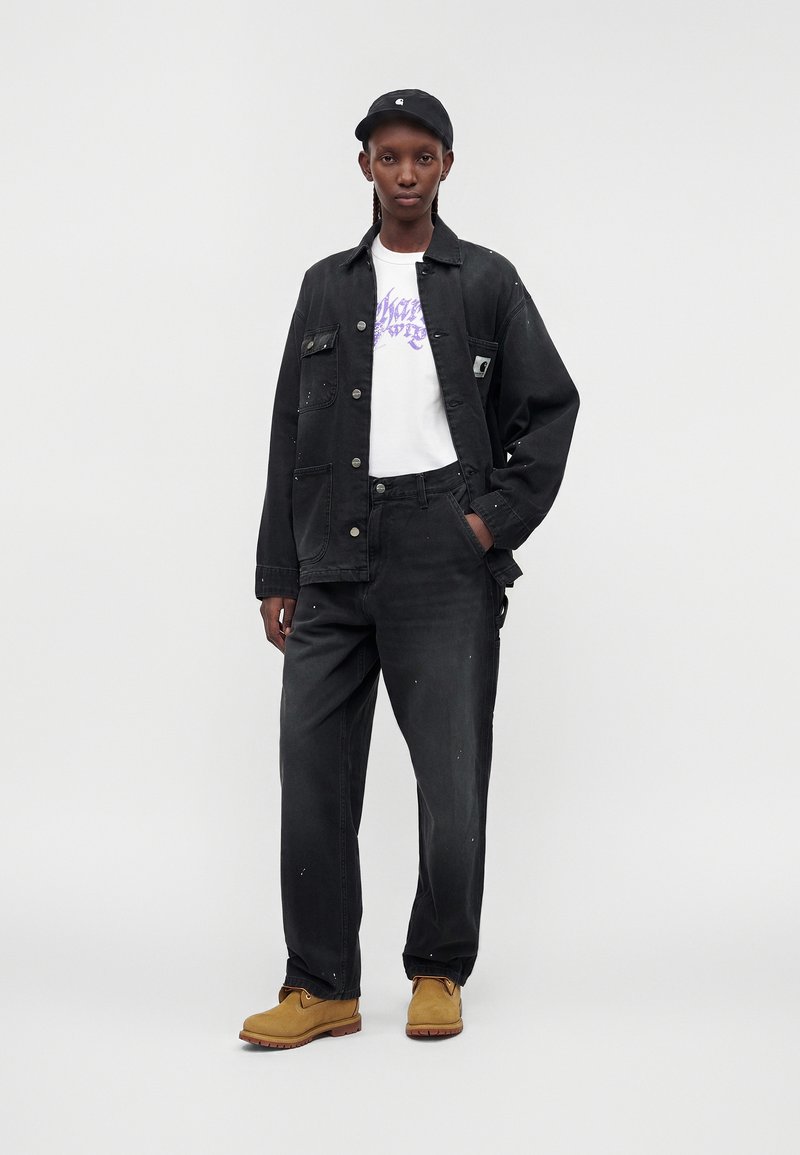 Stehende Person, die eine schwarze Mütze, eine schwarze Jacke, schwarze Hose, ein weißes T-Shirt mit Grafik und beige Arbeitsschuhe trägt, posiert vor einem einfarbigen hellen Hintergrund.