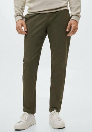 Homme portant un pantalon vert olive, une chemise beige à manches longues et des baskets blanches, debout devant un fond uni.