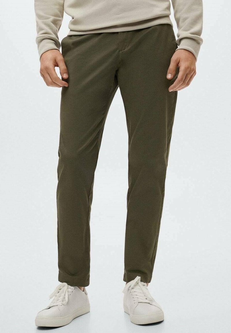Homme portant un pantalon vert olive, une chemise beige à manches longues et des baskets blanches, debout devant un fond uni.