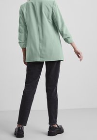 Mint green tailored blazer with three-quarter sleeves, paired with dark slim-fit trousers and black platform shoes. Smooth fabric, minimal design.