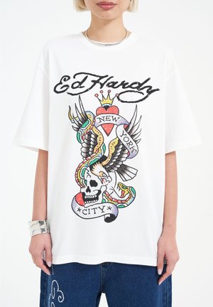 Bílý bavlněný tričko s odvážným, barevným tetovacím designem, který zahrnuje lebku, křídla, srdce a text - "Ed Hardy," "New York," "City."
