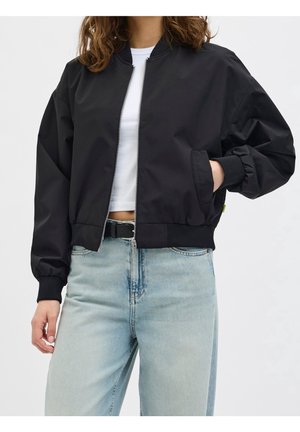 Person trägt eine schwarze Bomberjacke mit Reißverschluss vorne über einem weißen bauchfreien Top und hellblauen, hoch taillierten Jeans mit schwarzem Gürtel.