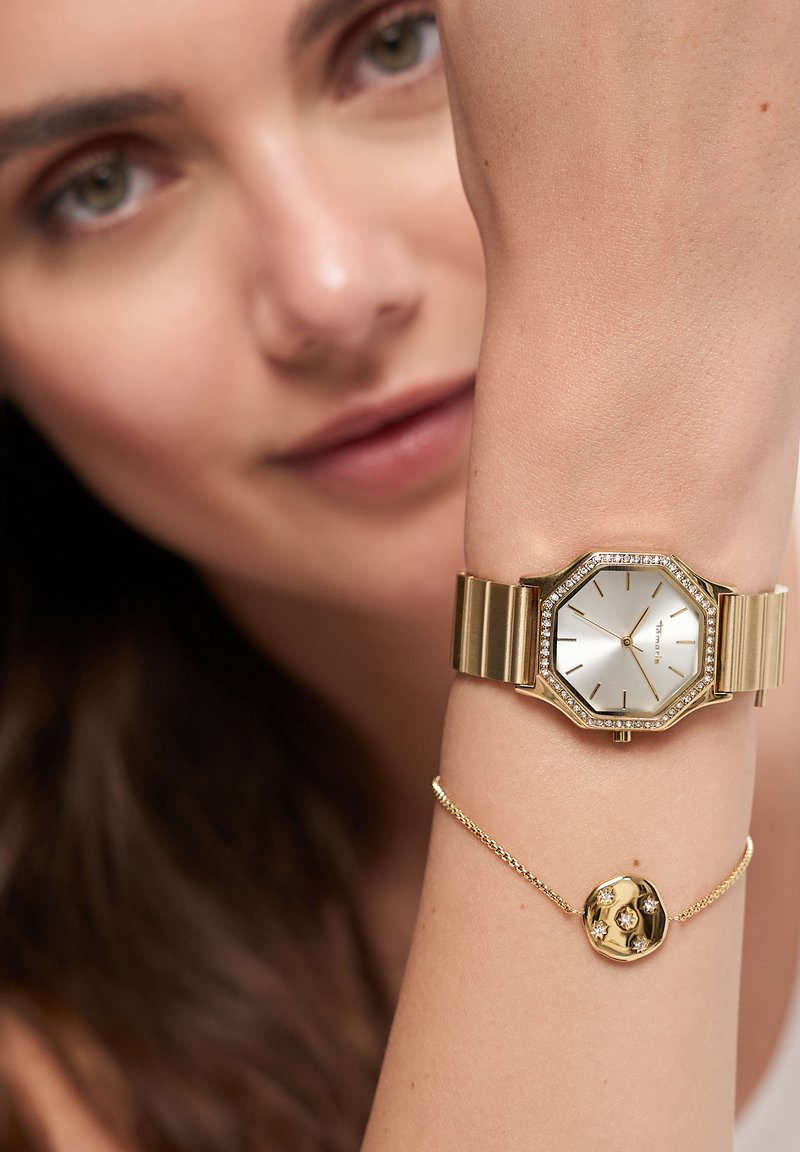 Guld octagonalt ur med en sølv ansigt og krystal udsmykninger, parret med et delikat guld armbånd med en rund charm og små sten.