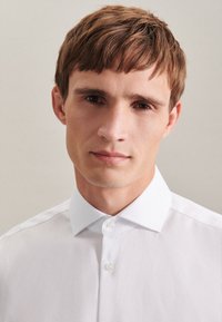 Camicia bianca con colletto classico, caratterizzata da una texture liscia e una silhouette aderente, indossata da un modello con capelli corti.