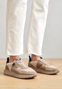Zapatillas beige con ante, detalles de piel texturizada, cierre de cordones y lengüetas en negro. Combínalas con jeans blancos con dobladillo deshilachado sobre un suelo de madera.