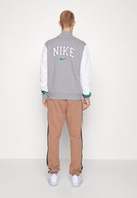 Giacca varsity grigia Nike con maniche bianche, dettagli verdi e logo ricamato; abbinata a pantaloni della tuta marroni con strisce laterali e scarpe bianche.