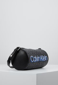 Borsa cilindrica nera con tessuto decorato con il logo "Calvin Klein" in blu. Include una tracolla nera staccabile e dettagli in pelle.
