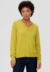 V-Ausschnitt Pullover in einem lebhaften Limettengrün mit Strickstruktur, langen Ärmeln, gerippten Bündchen und Saum, entspannte Passform. Kombiniert mit dunklen Jeans.