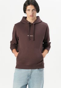 Brown cotton hoodie with a drawstring hood, front kangaroo pocket, ribbed cuffs, and embroidered text on the chest. Worn with light blue jeans.