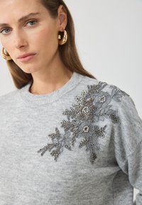 Sudadera gris con un intrincado diseño floral de cuentas y bordado en un hombro. Textura suave con cuello redondo clásico.