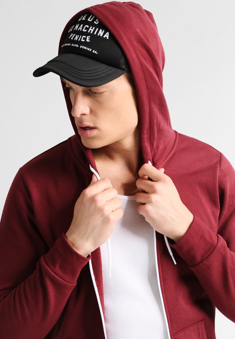 Sudadera burdeos con cremallera y cordones, usada sobre una camiseta de tirantes blanca de canalé. Una gorra negra con texto blanco completa el atuendo.