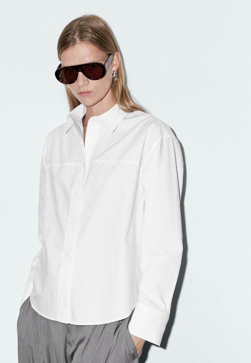 Chemise blanche en coton avec un col pointu, des manches longues et une fermeture à boutons. Complétée par des lunettes de soleil ovales sombres et un pantalon gris à plis.