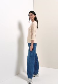 Femme debout de profil portant un haut beige à manches longues, un jean bleu à jambes larges et des baskets argentées, sur un fond clair uni.