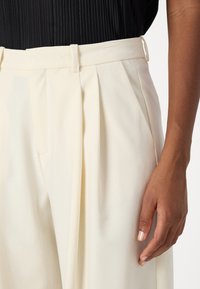 Cream pleated trousers with a tailored fit, featuring belt loops and a smooth, textured surface. Hand partially visible on the side.