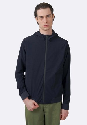 Uomo che indossa una giacca nera con cappuccio e cerniera e pantaloni verdi, in piedi davanti a uno sfondo grigio chiaro uniforme.