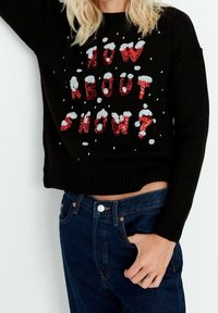 Henkilö, jolla on päällään musta villapaita, jossa on punaisella ja valkoisella paljetti-tekstillä "Mitä sanot lumesta?", sekä tummansiniset farkut, käsi taskussa.