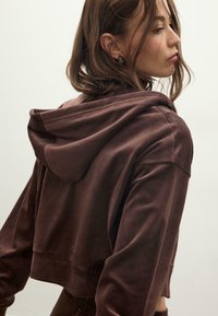 Brązowy krótki hoodie wykonany z miękkiego materiału, z kieszenią kangurzą i kapturem na sznurku. Posiada luźny krój oraz ściągacze przy mankietach.