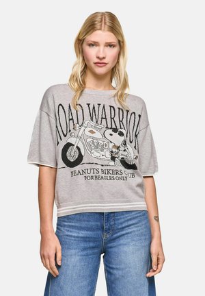 Woman with long blonde hair wears a gray “Road Warrior” Snoopy biker t-shirt and blue jeans, standing against a plain white background.