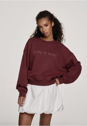 Donna con capelli ricci che indossa una felpa color melograno con la scritta "GIRL'S GIRL" e una gonna bianca a bolle, posando davanti a uno sfondo semplice.