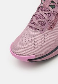 Baskets de sport rose clair avec une tige en tissu texturé, bout renforcé et semelle foncée contrastante. Présente un design à lacets et des trous de ventilation.