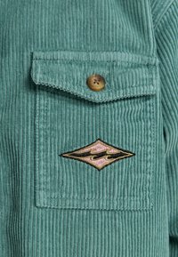 Camicia in corduroy color teal con taschino sul petto dotato di bottone e un logo a diamanté ricamato in nero, rosa e giallo. Texture verticale a righe.