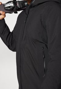 Osoba v černé zipové bundě drží lyžařské vázání na svém rameni před jednobarevným pozadím.