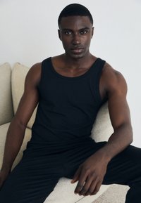 Sort tanktop og matchende bukser, afslappet pasform, glat tekstur, båret af en person, der sidder på en lysefarvet sofa. Simpelt design.