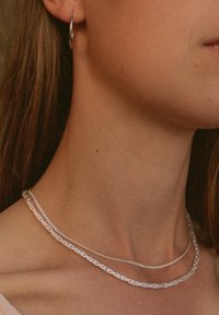 Silberne Halsketten übereinander am Hals getragen; eine mit einem Ketten-Design, die andere in einem glatten, dünnen Stil. Runde silberne Creolen sind sichtbar.