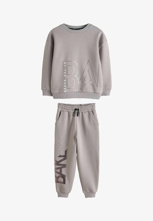 Teenager steht mit Händen in den Taschen, trägt einen grauen übergroßen Sweatshirt und passende Sweatpants mit weißen Sneakers vor neutralem Hintergrund.