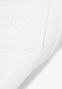 Gros plan sur un tissu blanc texturé avec le logo "LACOSTE PARIS" en relief embossé sur la surface.
