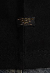 Etiqueta de tela negra con texto dorado que dice "LIBERTAD ILIMITADA", "PROBADO A GRAN ALTITUD", y "TIPO N° C-46." Diseño sencillo, forma rectangular.