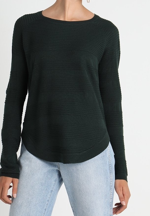 Femme portant un pull texturé vert foncé à manches longues avec un jean bleu clair sur un fond blanc uni.
