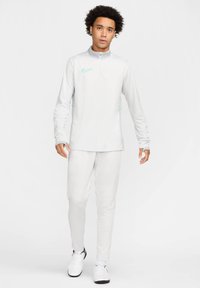 Weißes Nike Zip-Up Langarmoberteil und passende Hose. Hellblaue Akzente auf dem Oberteil. Glatte Textur. Das Modell trägt weiße Sportschuhe.