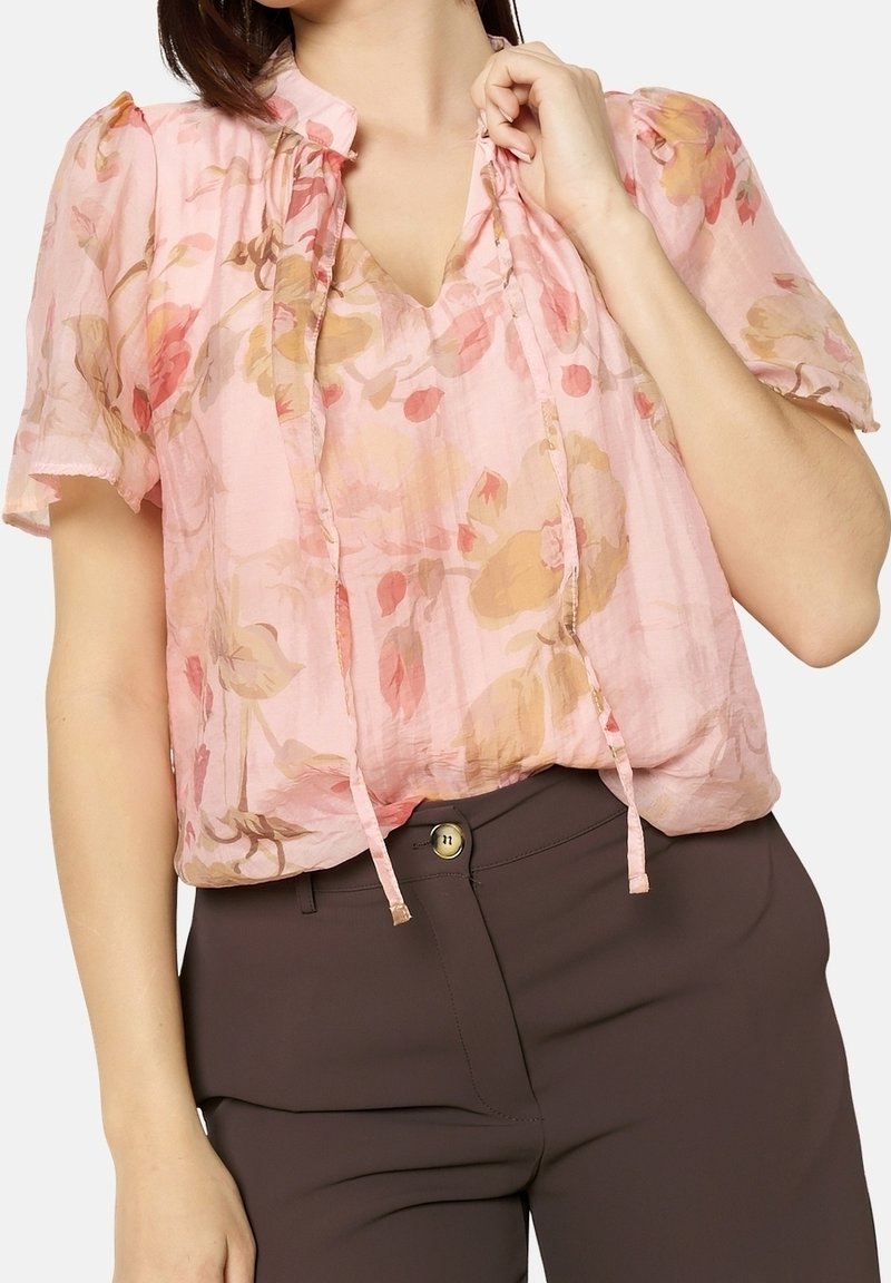 Blouse fleurie en chiffon rose pâle avec manches bouffantes et décolleté en V, ornée d'un nœud au col et de motifs floraux pastel doux.