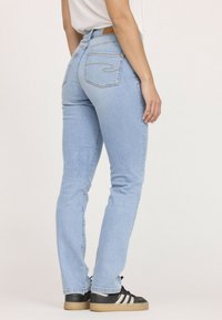 Jean en denim bleu clair avec une taille haute, coupe slim et deux poches arrière. Porté avec des chaussures de sport noires avec des rayures blanches.