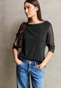 Haut en tissu imprimé léopard noir à manches longues, associé à un jean en denim bleu clair. Le mannequin pose avec assurance, les mains dans les poches. Sac marron en accent.