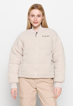 Donna con capelli biondi lisci che indossa una giacca in pile Columbia beige chiaro e pantaloni cargo beige, in piedi su uno sfondo bianco.