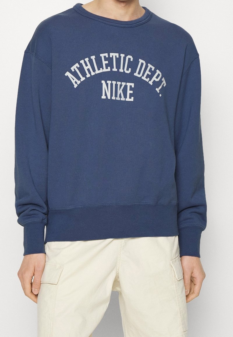 Homme portant un sweat-shirt bleu marine avec "ATHLETIC DEPT. NIKE" en lettres blanches et un pantalon cargo beige clair.