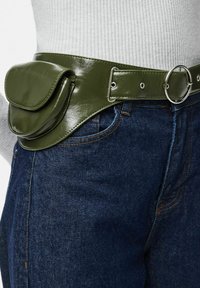 Zelený, lesklý pásek z umělé kůže s kapsou, stříbrnými kovovými prvky, nosí se přes modré džíny. Má sponu a hladký povrch.