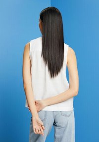 Weiße ärmellose Bluse mit lockerem Schnitt, kombiniert mit hellblauen Jeans. Die Frisur ist lang und glatt, vor einem blauen Hintergrund.