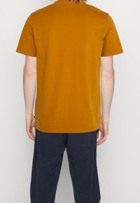 Musztardowy t-shirt z krótkim rękawem wykonany z bawełny, z klasycznym okrągłym dekoltem i prostym wykończeniem, zestawiony z granatowymi spodniami.