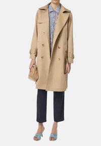 Trench coat beige con design doppiopetto, bottoni dorati e spalline, abbinato a pantaloni scuri e sandali con tacco blu.