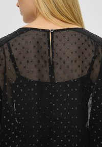 Schwarzes transparentes Oberteil aus strukturiertem Stoff mit kleinen glitzernden Punkten. Knopfverschluss hinten, mit einem Schlitzdetail direkt unterhalb des Ausschnitts.