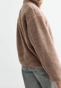 Ruskea fleece-takki, jossa on korkea kaulus ja karhea, pörröinen pinta. Löysä istuvuus, näkyvät saumat ja hieman pyöristetty helma.