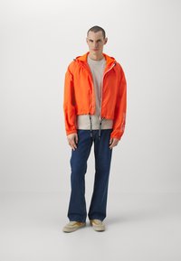 Afkortet orange jakke med hætte, elastiske manchetter og snoretræk; båret over en beige skjorte med blå jeans og sko med gule accenter.